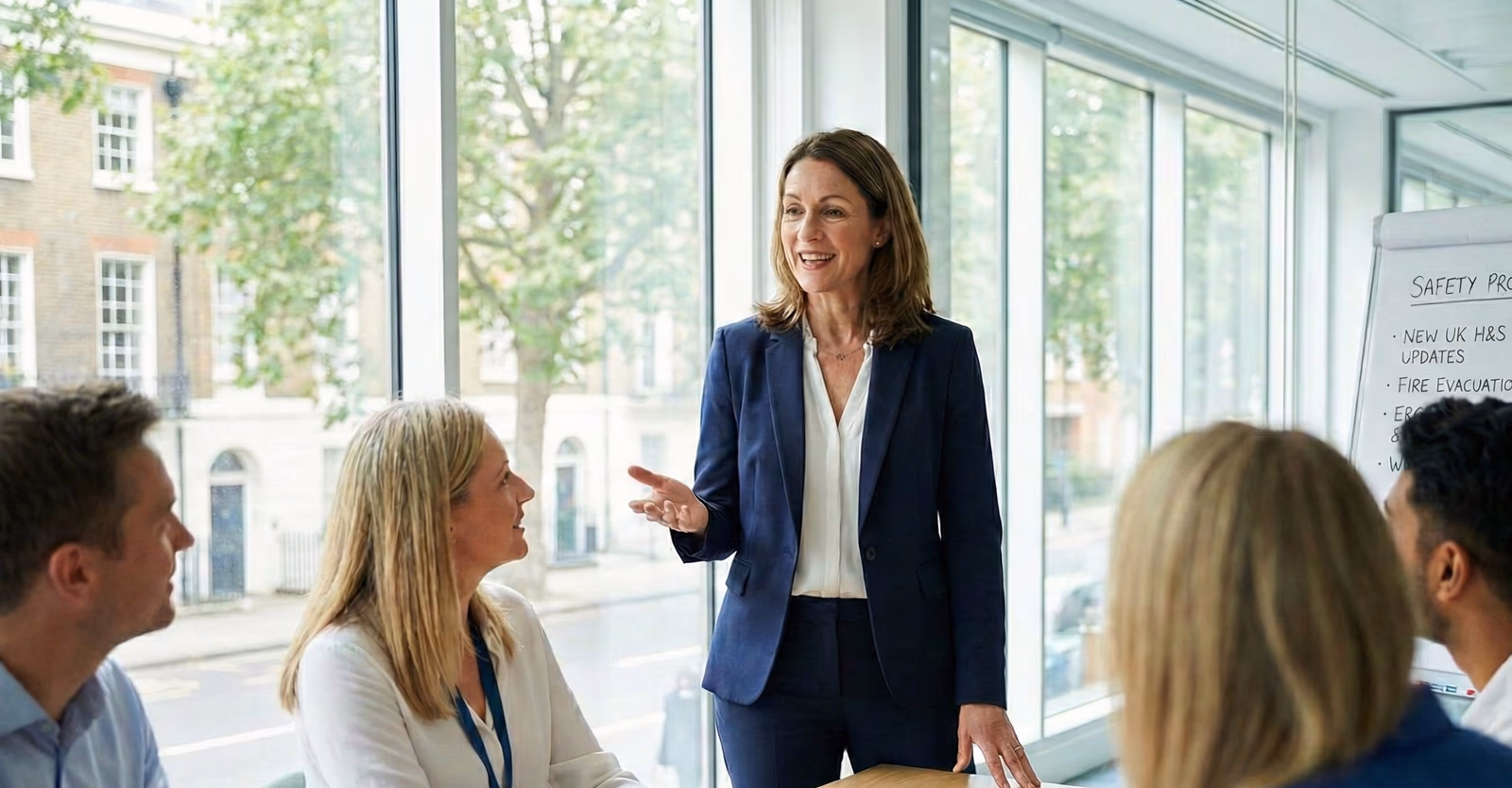 A safety professional leading a team briefing in a modern office, with a whiteboard showing health and safety agenda items including UK H&S updates and fire evacuation procedures.